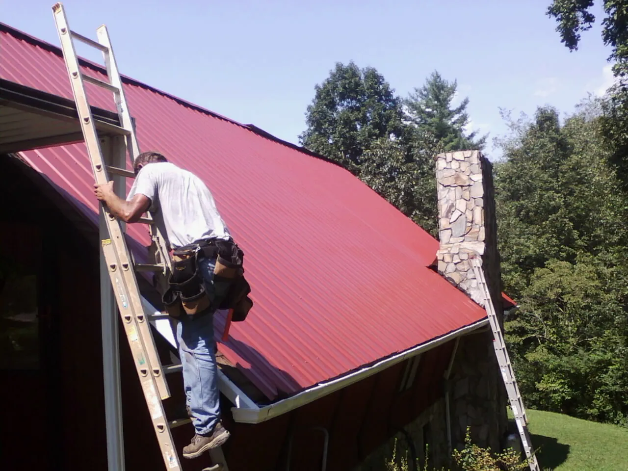 Roofer on a ladder beside a red metal roof and stone chimney — Soffit & Fascia Repair by Roofing Brothers in Daykin